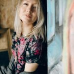 A stylish blonde woman poses against colorful artwork, exuding confidence and elegance.