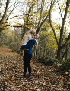 A woman joyfully embraces her dog on a foliage-covered forest trail in autumn.